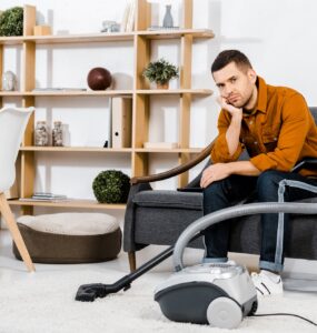 upset man in modern living room sitting on sofa near hoover and looking at camera