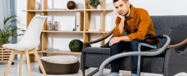 upset man in modern living room sitting on sofa near hoover and looking at camera