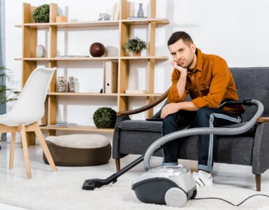 upset man in modern living room sitting on sofa near hoover and looking at camera